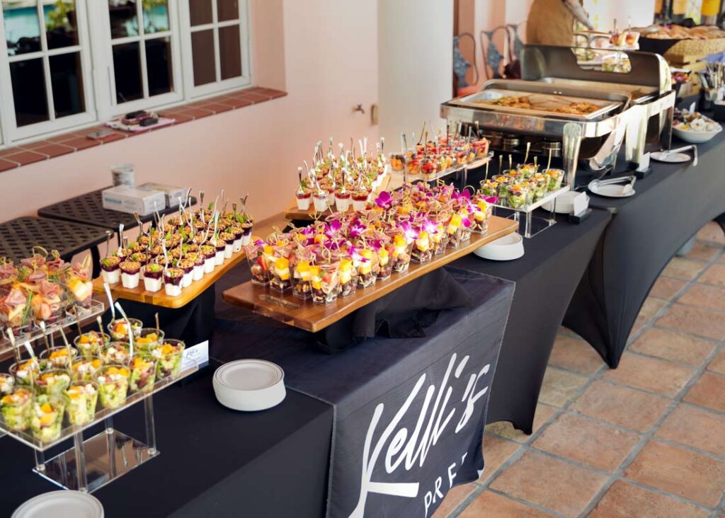 Elegant buffet table by Kelli’s Catering featuring colorful individual appetizer cups, fresh fruit parfaits with orchids, and hot entrées in chafing dishes, set up for an outdoor event